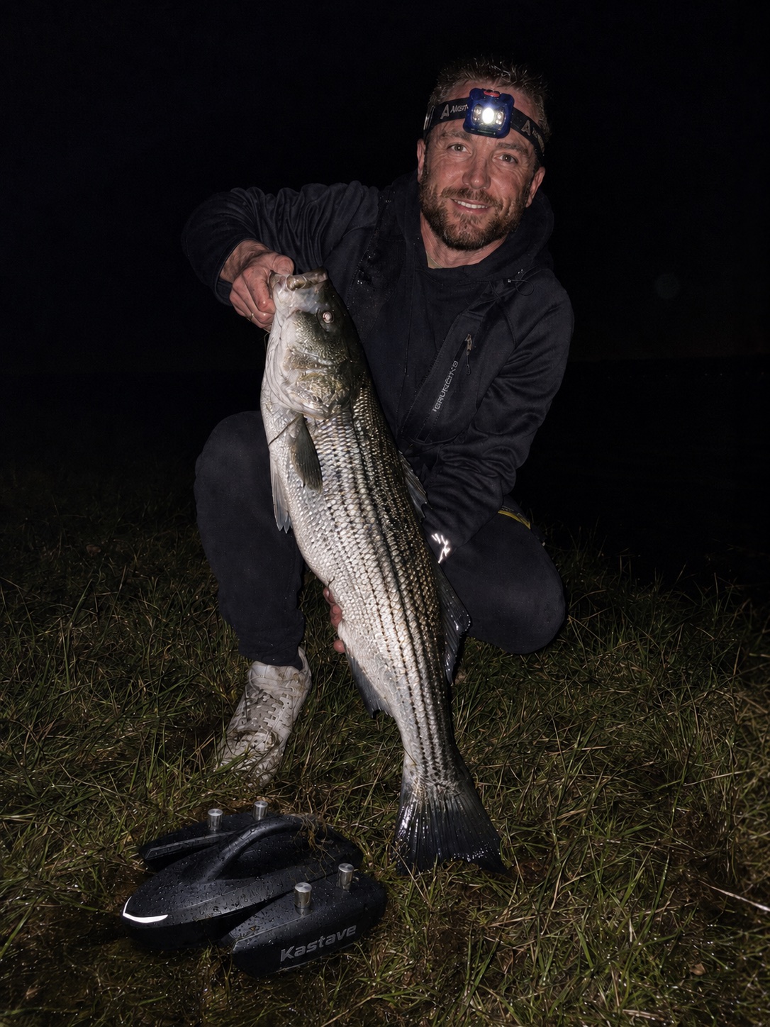 Early tester holding a striped bass at night with Kastave nearby